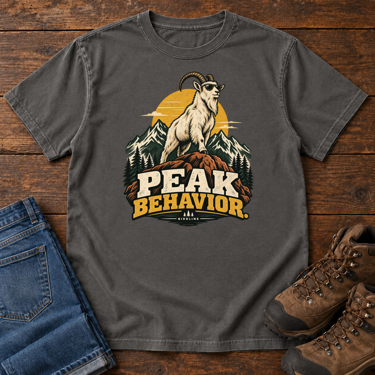 Peak Behavior T-Shirt in Pepper, front laid flat on wood, featuring a horned white goat wearing sunglasses on a rocky peak, with mountains, a gold sun and bold lettering.