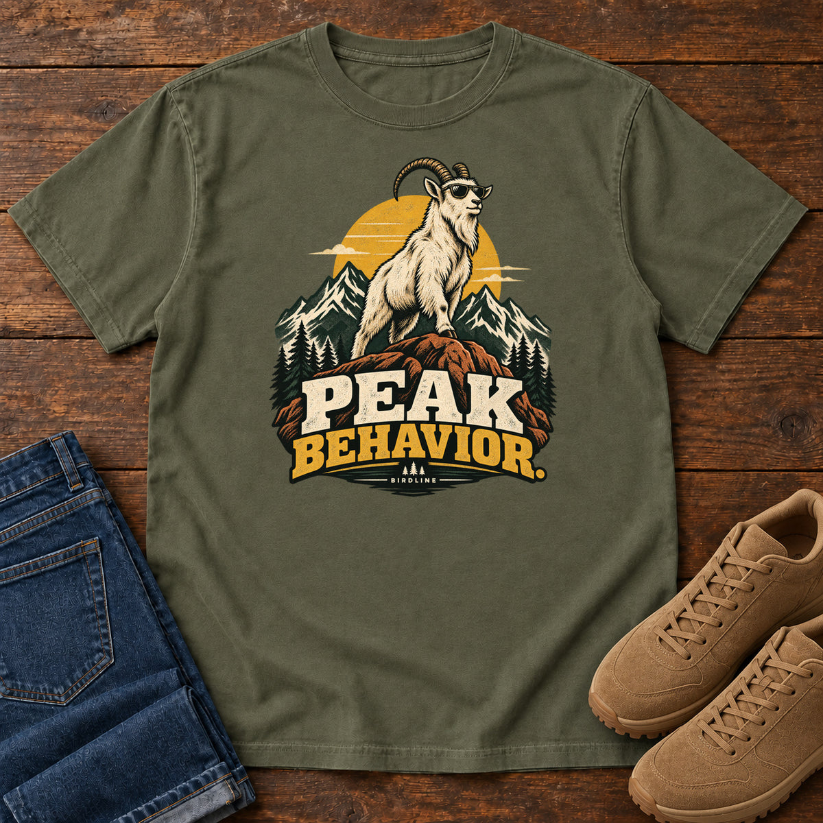 Peak Behavior T-Shirt in Sage, front laid flat on wood, featuring a horned white goat wearing sunglasses on a rocky peak, with mountains, a gold sun and bold lettering.