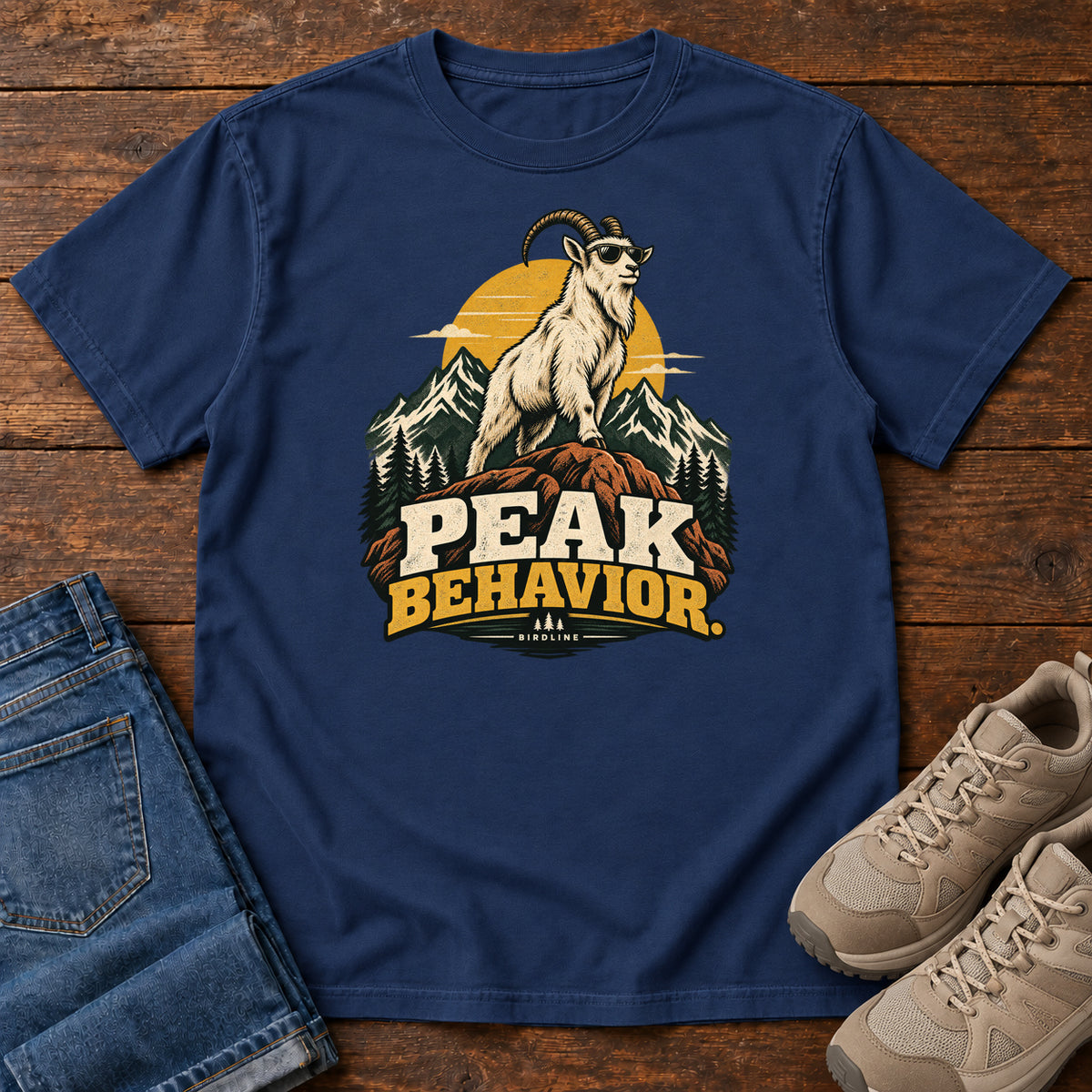Peak Behavior T-Shirt in True Navy, front laid flat on wood, featuring a horned white goat wearing sunglasses on a rocky peak, with mountains, a gold sun and bold lettering.
