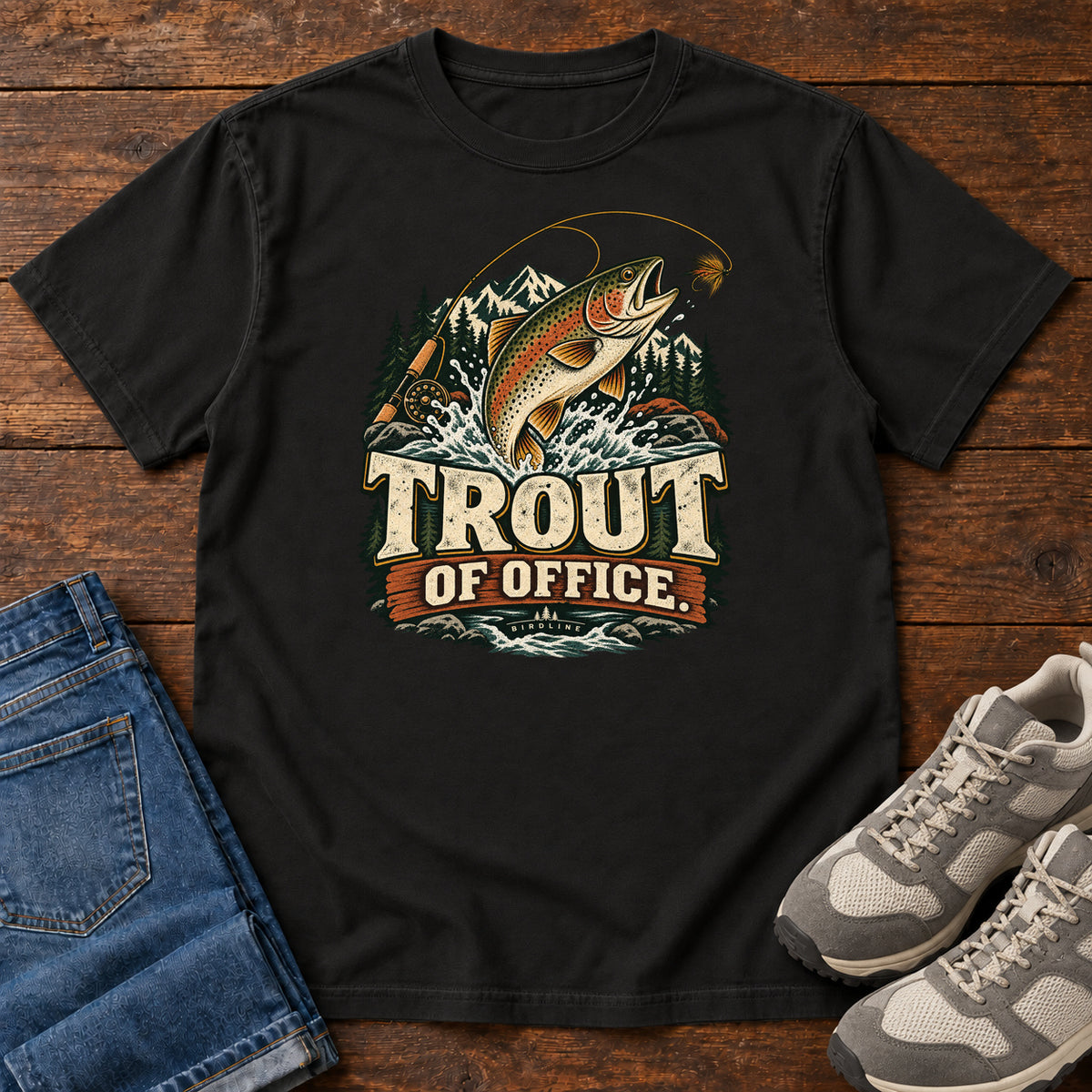 Black Trout of Office Fly Rod T-Shirt with a spotted fish leaping toward a fishing fly, with mountains and cream lettering, front flat lay on wood.