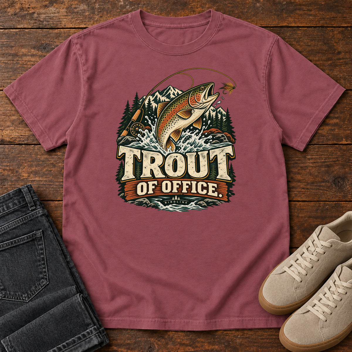 Brick Trout of Office Fly Rod T-Shirt with a spotted fish leaping toward a fishing fly, with mountains and cream lettering, front flat lay on wood.