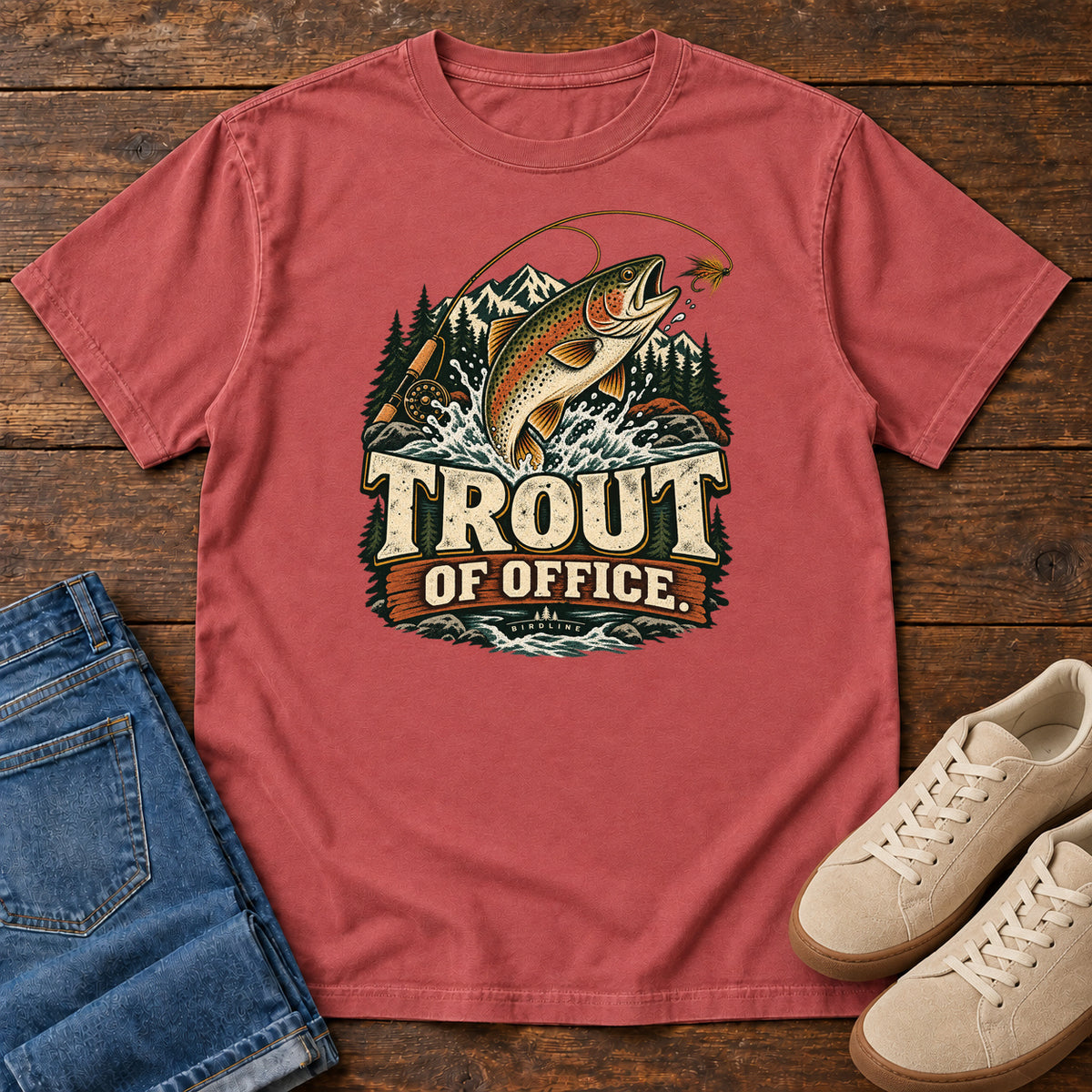 Crimson Trout of Office Fly Rod T-Shirt with a spotted fish leaping toward a fishing fly, with mountains and cream lettering, front flat lay on wood.