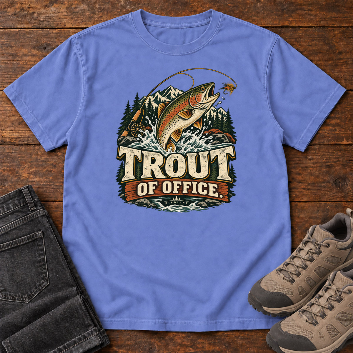 Flo Blue Trout of Office Fly Rod T-Shirt with a spotted fish leaping toward a fishing fly, with mountains and cream lettering, front flat lay on wood.