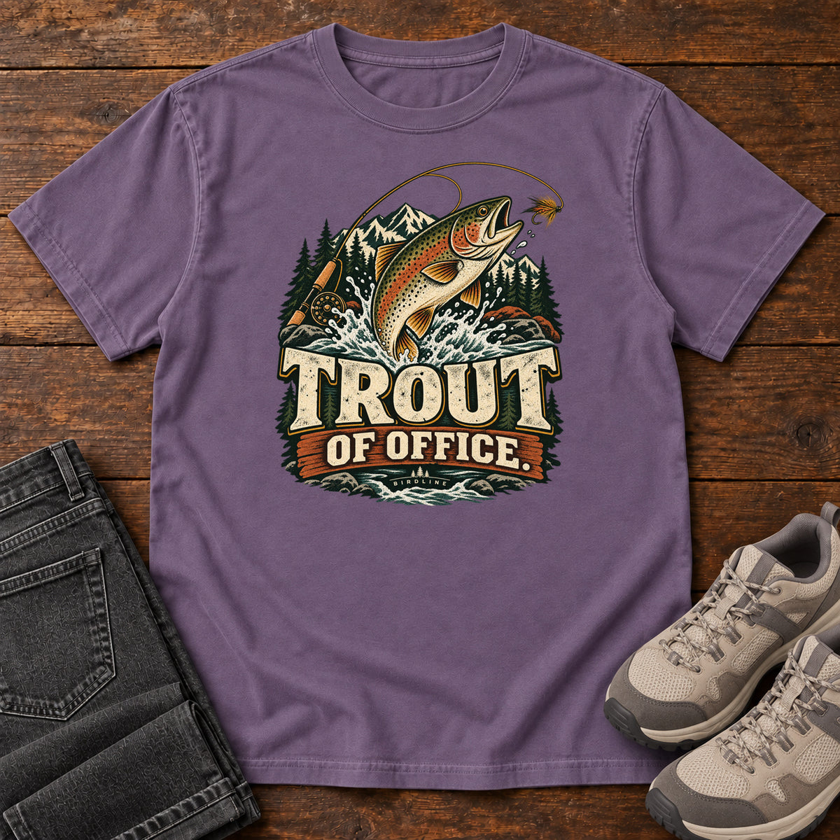 Grape Trout of Office Fly Rod T-Shirt with a spotted fish leaping toward a fishing fly, with mountains and cream lettering, front flat lay on wood.