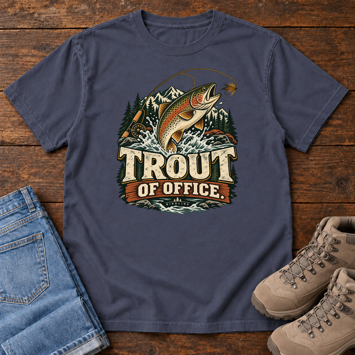 Navy Trout of Office Fly Rod T-Shirt with a spotted fish leaping toward a fishing fly, with mountains and cream lettering, front flat lay on wood.