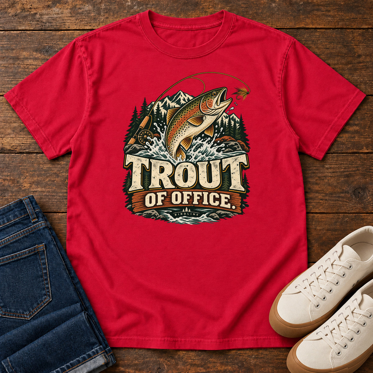 Red Trout of Office Fly Rod T-Shirt with a spotted fish leaping toward a fishing fly, with mountains and cream lettering, front flat lay on wood.