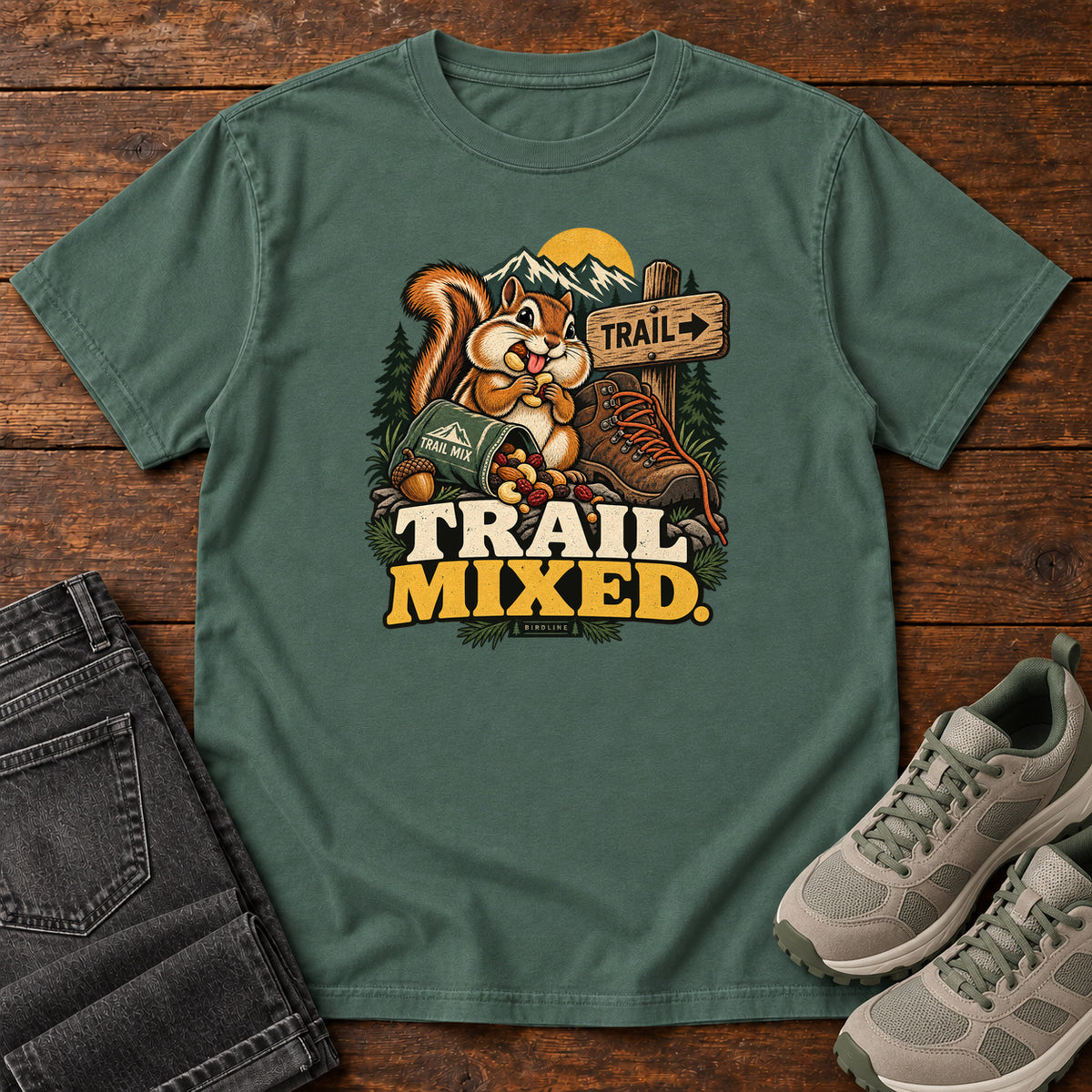 Blue Spruce Trail Mixed T-Shirt with a bushy-tailed cartoon animal beside a trail-mix bag, hiking boot and trail sign, front flat lay on wood.