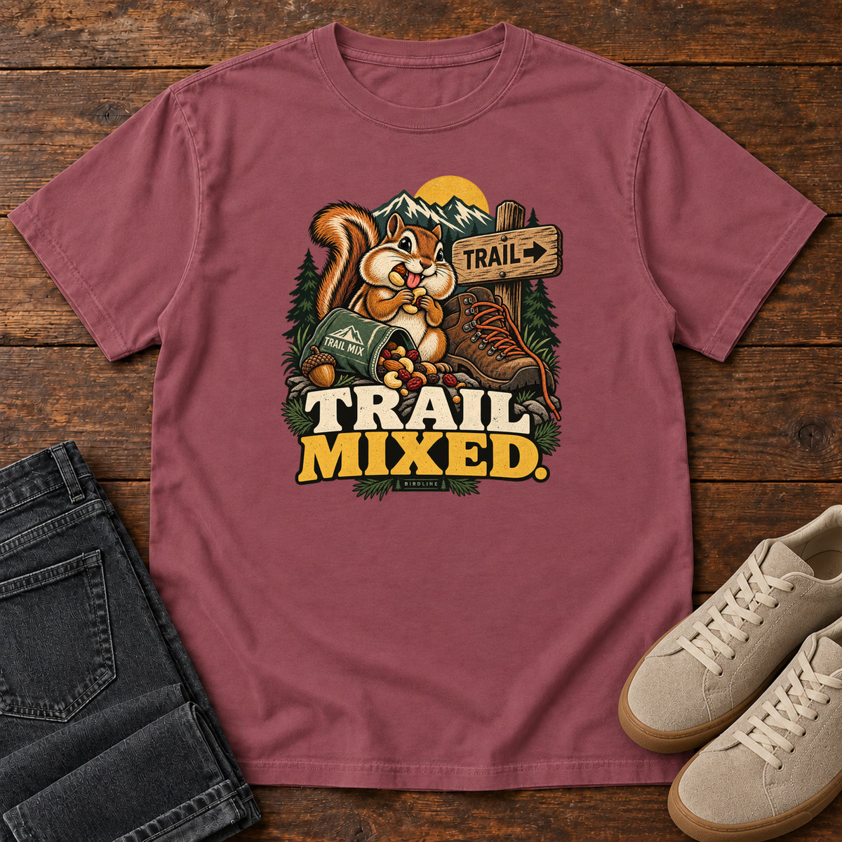 Brick Trail Mixed T-Shirt with a bushy-tailed cartoon animal beside a trail-mix bag, hiking boot and trail sign, front flat lay on wood.