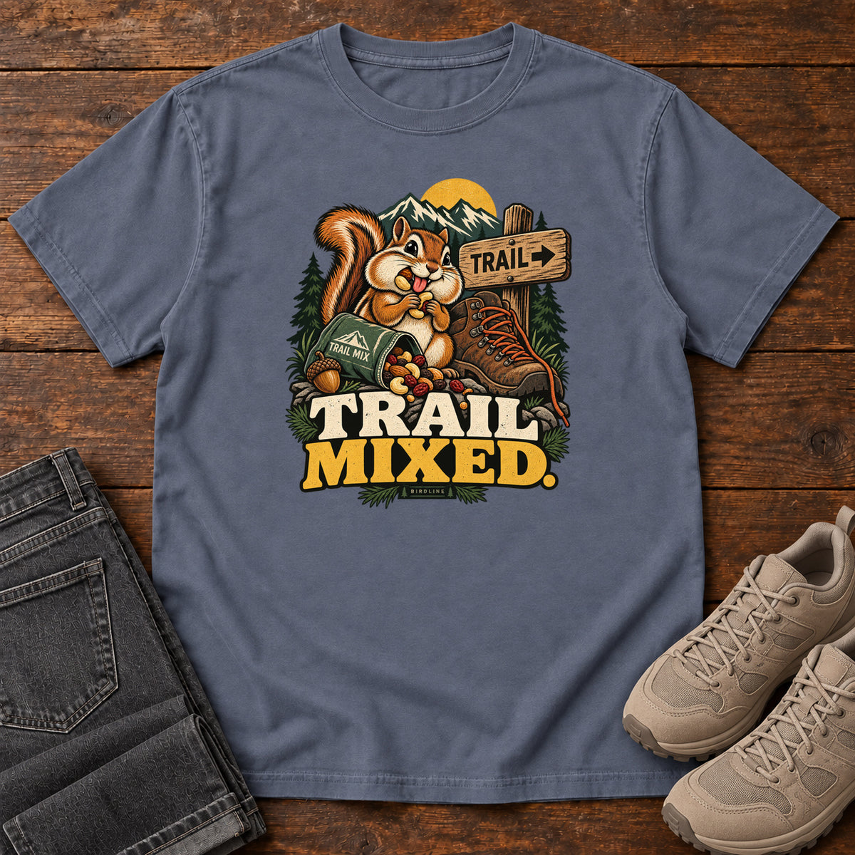 Denim Trail Mixed T-Shirt with a bushy-tailed cartoon animal beside a trail-mix bag, hiking boot and trail sign, front flat lay on wood.
