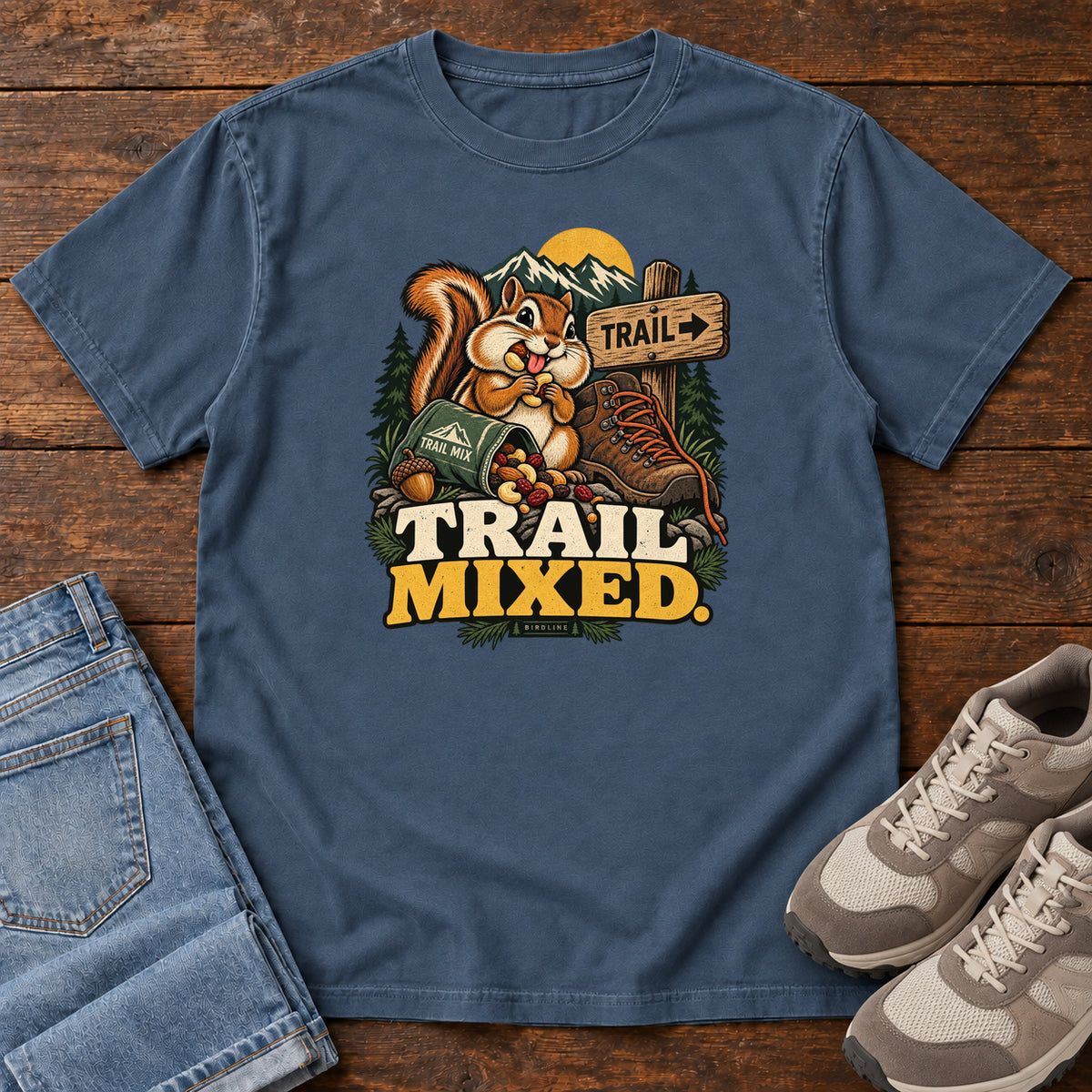 Midnight Trail Mixed T-Shirt with a bushy-tailed cartoon animal beside a trail-mix bag, hiking boot and trail sign, front flat lay on wood.