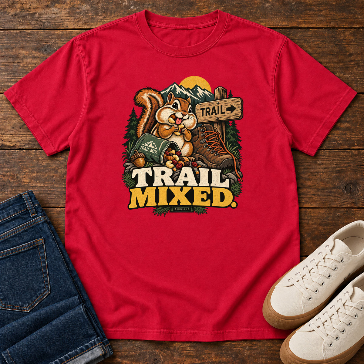 Red Trail Mixed T-Shirt with a bushy-tailed cartoon animal beside a trail-mix bag, hiking boot and trail sign, front flat lay on wood.
