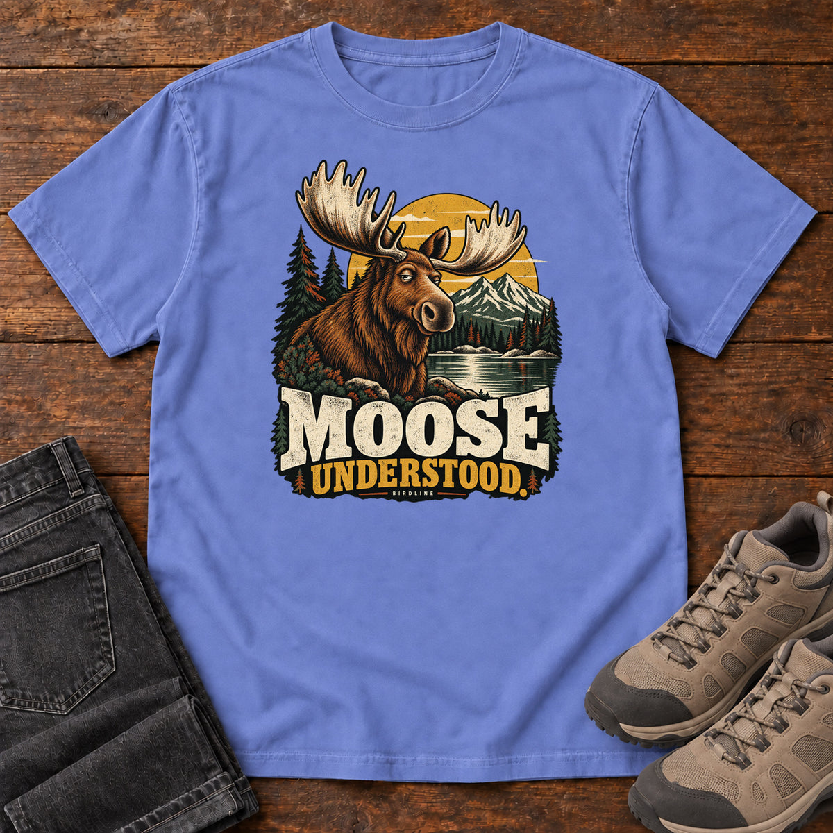 Flo Blue Moose Understood T-Shirt with an antlered moose beside a mountain lake, pines and a golden sun; front flat lay on wood.
