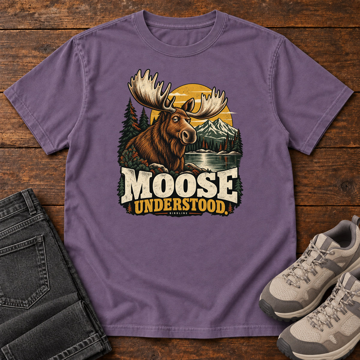 Grape Moose Understood T-Shirt with an antlered moose beside a mountain lake, pines and a golden sun; front flat lay on wood.