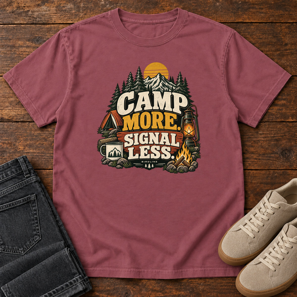 Brick Camp More Signal Less T-Shirt with a tent, lantern, campfire and mountain scene around bold slogan lettering; front flat lay on wood.