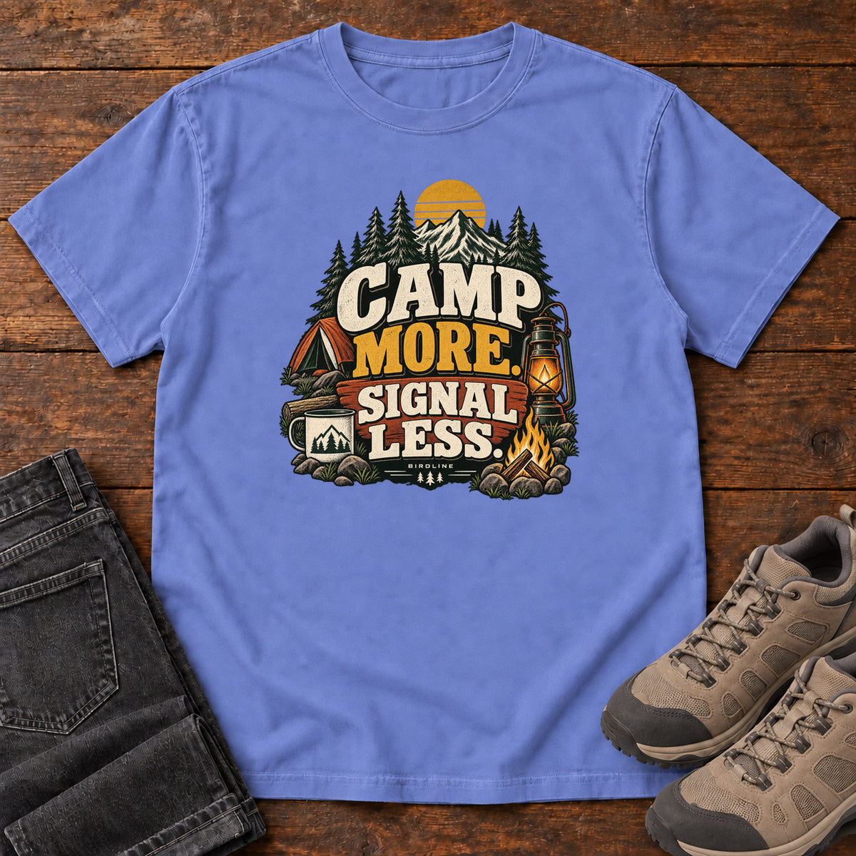 Flo Blue Camp More Signal Less T-Shirt with a tent, lantern, campfire and mountain scene around bold slogan lettering; front flat lay on wood.
