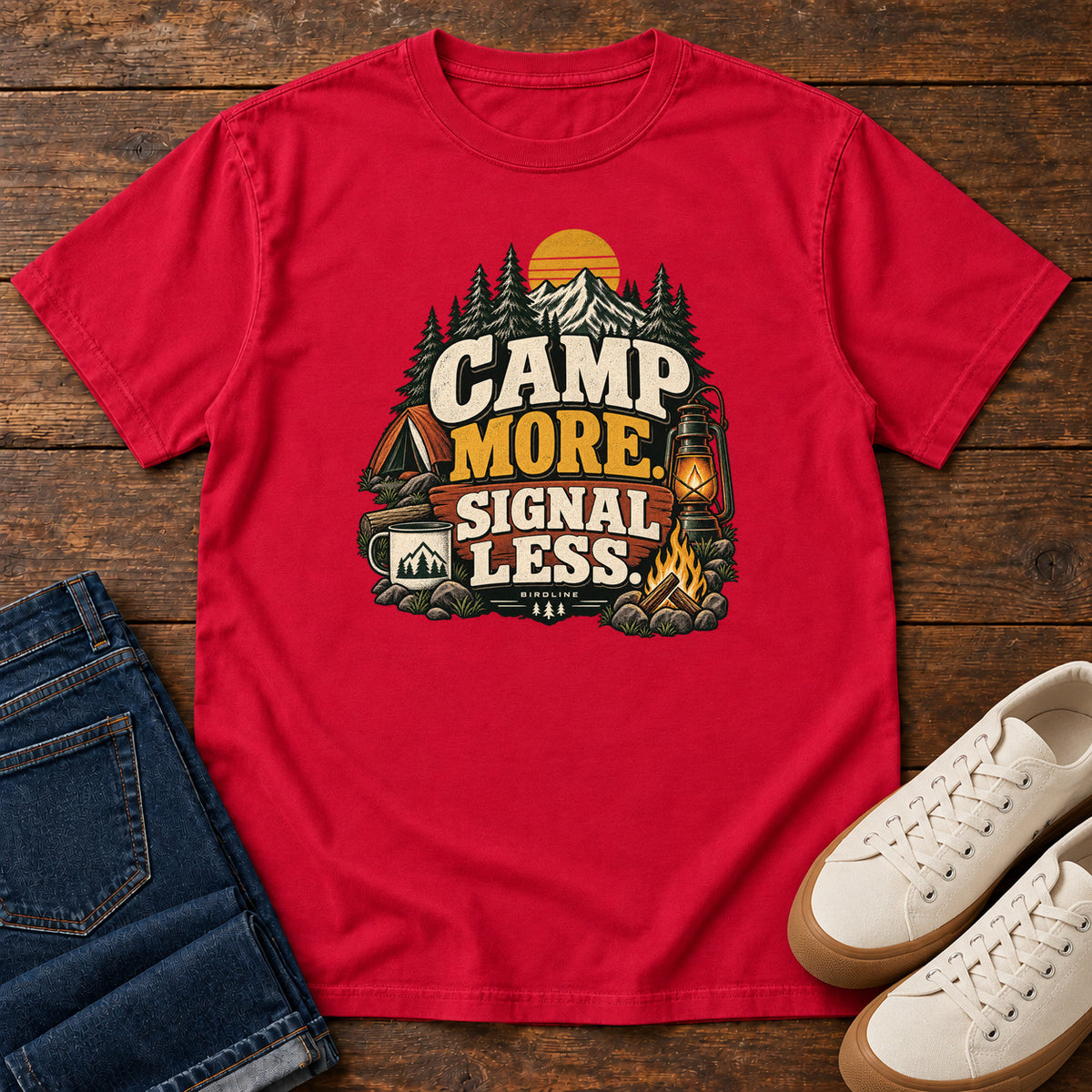 Red Camp More Signal Less T-Shirt with a tent, lantern, campfire and mountain scene around bold slogan lettering; front flat lay on wood.