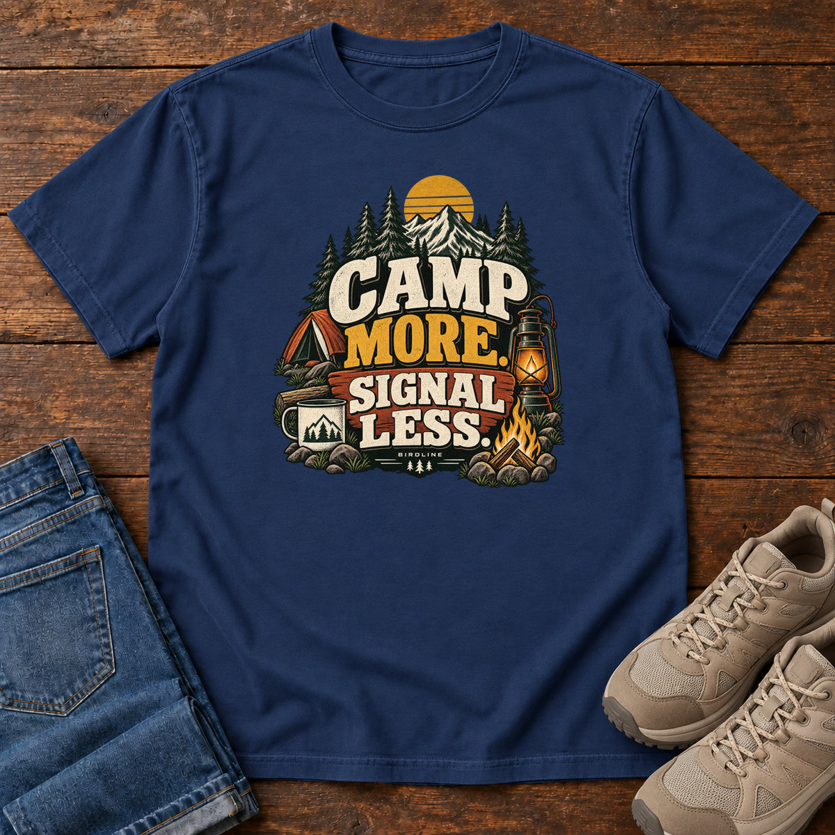True Navy Camp More Signal Less T-Shirt with a tent, lantern, campfire and mountain scene around bold slogan lettering; front flat lay on wood.