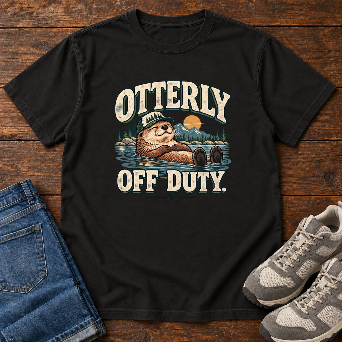 Black Otterly Off Duty T-Shirt with a floating otter in a knit cap, lake scenery and cream lettering, front flat lay on wood.