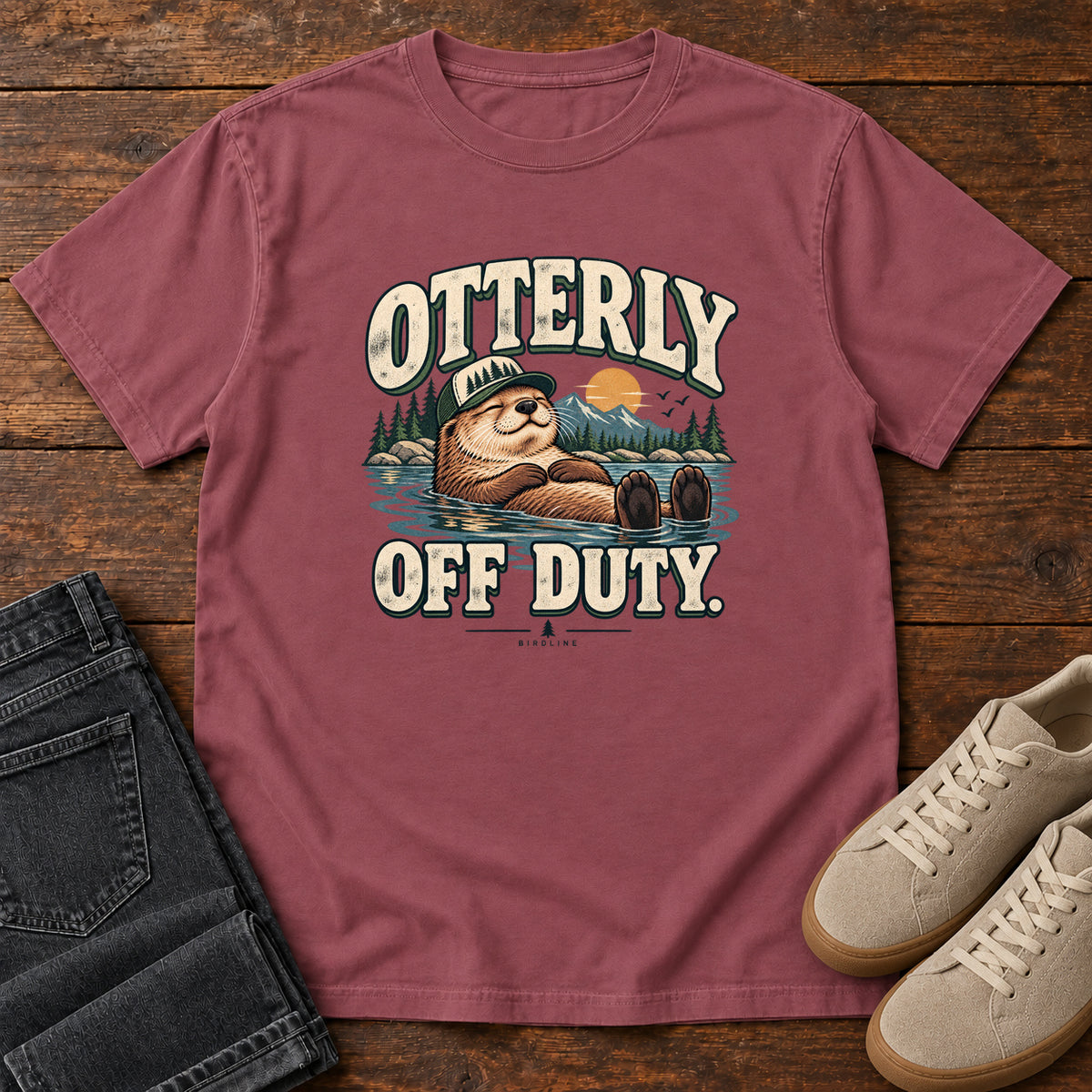 Brick Otterly Off Duty T-Shirt with a floating otter in a knit cap, lake scenery and cream lettering, front flat lay on wood.
