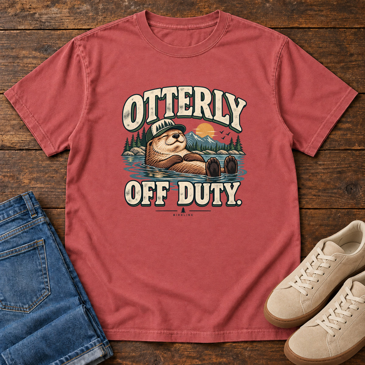 Crimson Otterly Off Duty T-Shirt with a floating otter in a knit cap, lake scenery and cream lettering, front flat lay on wood.