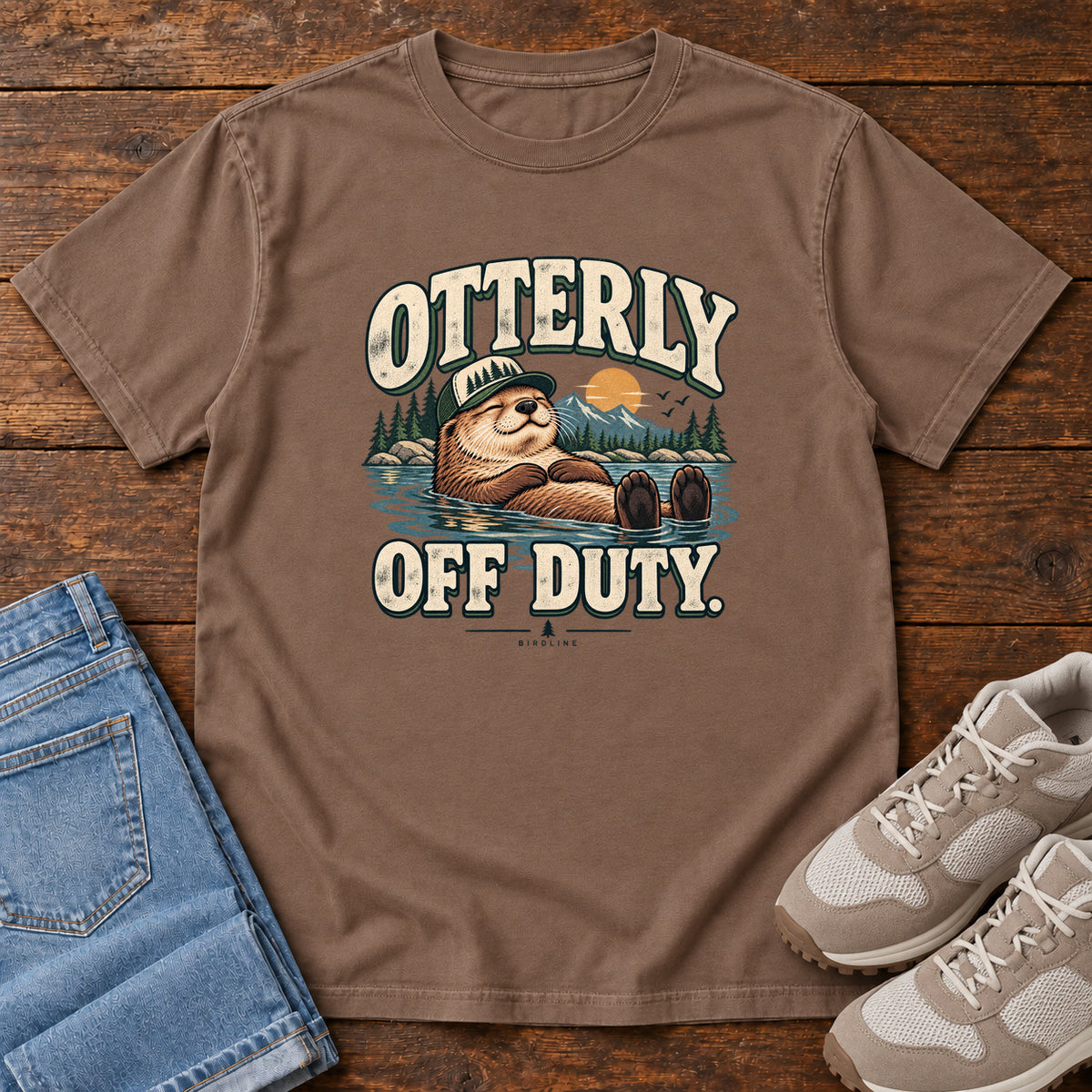 Espresso Otterly Off Duty T-Shirt with a floating otter in a knit cap, lake scenery and cream lettering, front flat lay on wood.