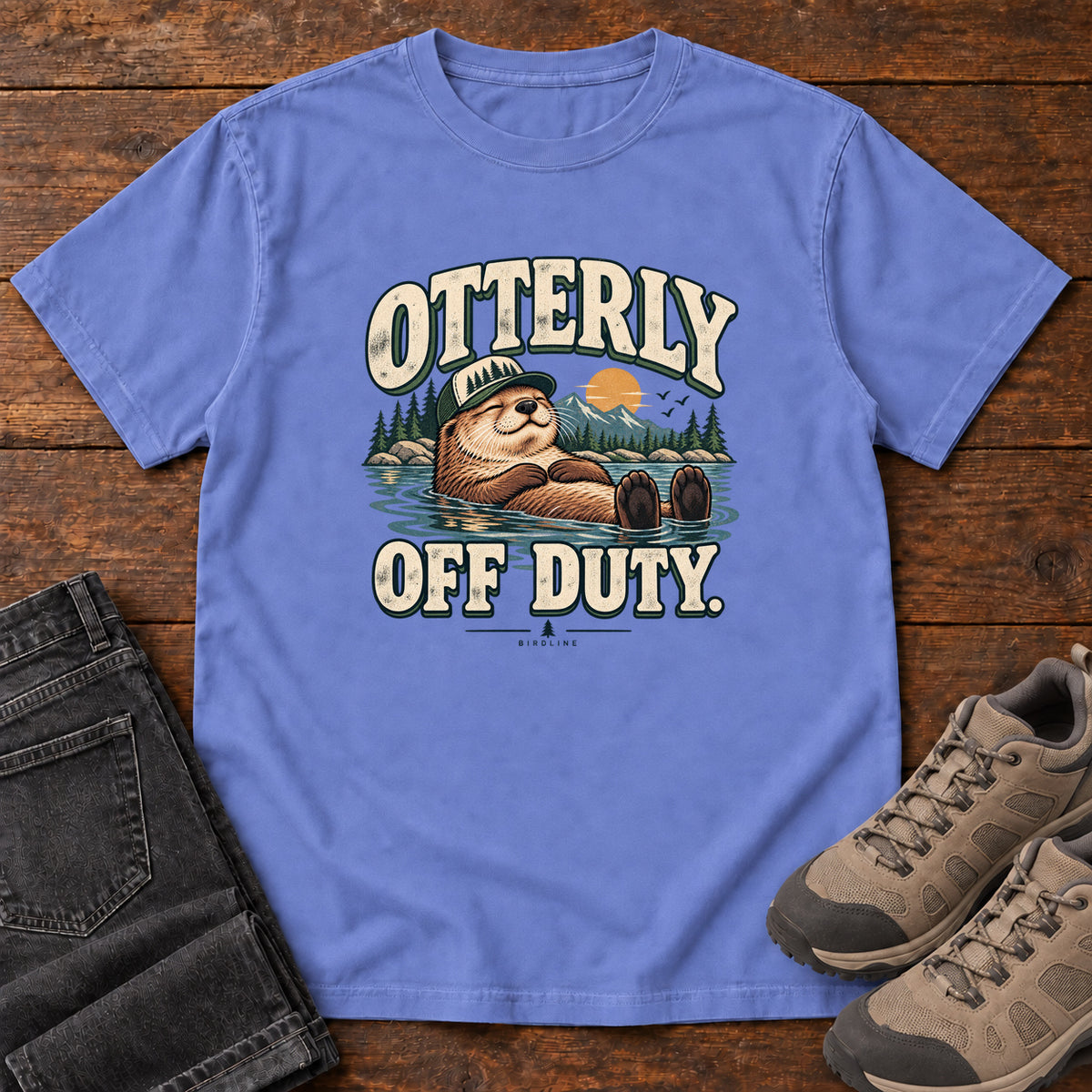 Flo Blue Otterly Off Duty T-Shirt with a floating otter in a knit cap, lake scenery and cream lettering, front flat lay on wood.