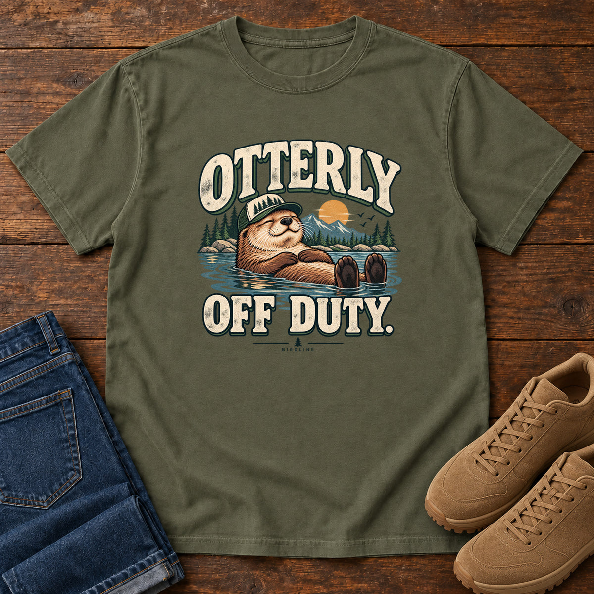 Sage Otterly Off Duty T-Shirt with a floating otter in a knit cap, lake scenery and cream lettering, front flat lay on wood.