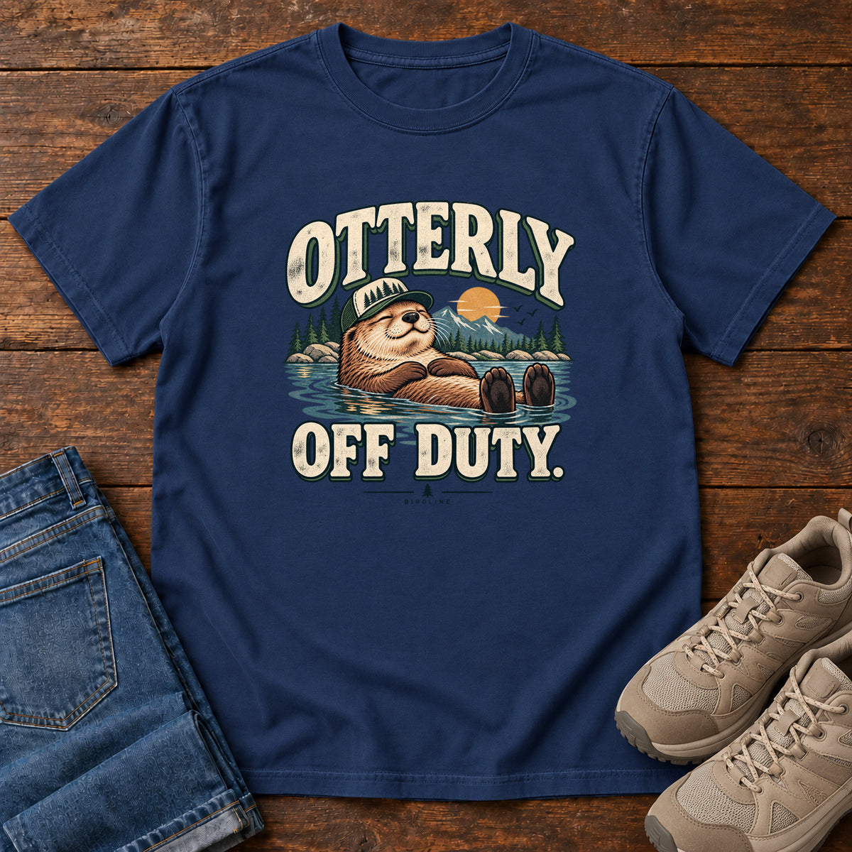 True Navy Otterly Off Duty T-Shirt with a floating otter in a knit cap, lake scenery and cream lettering, front flat lay on wood.