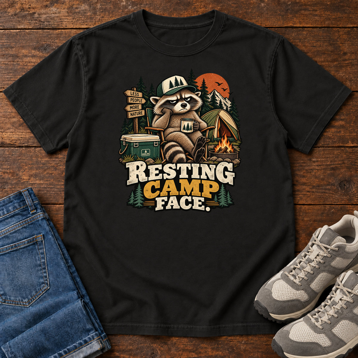 Resting Camp Face T-Shirt in Black, front laid flat on wood, featuring a capped raccoon resting in a camp chair with a mug beside a tent, cooler and campfire.