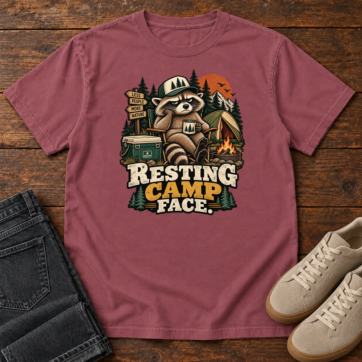 Resting Camp Face T-Shirt in Brick, front laid flat on wood, featuring a capped raccoon resting in a camp chair with a mug beside a tent, cooler and campfire.