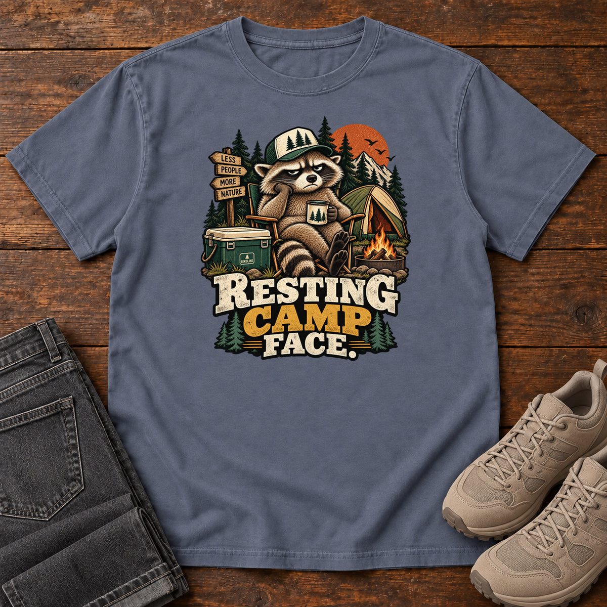 Resting Camp Face T-Shirt in Denim, front laid flat on wood, featuring a capped raccoon resting in a camp chair with a mug beside a tent, cooler and campfire.