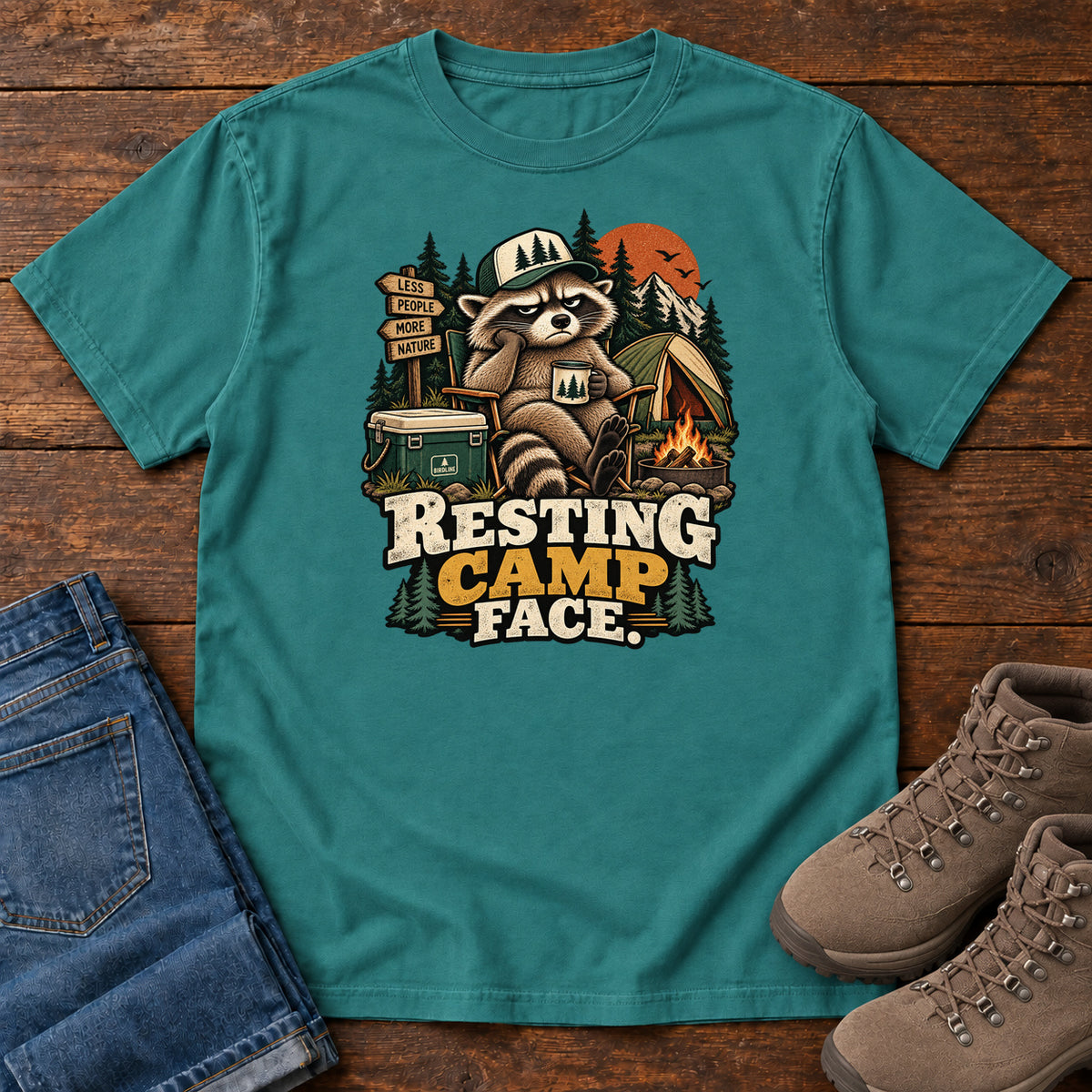 Resting Camp Face T-Shirt in Emerald, front laid flat on wood, featuring a capped raccoon resting in a camp chair with a mug beside a tent, cooler and campfire.