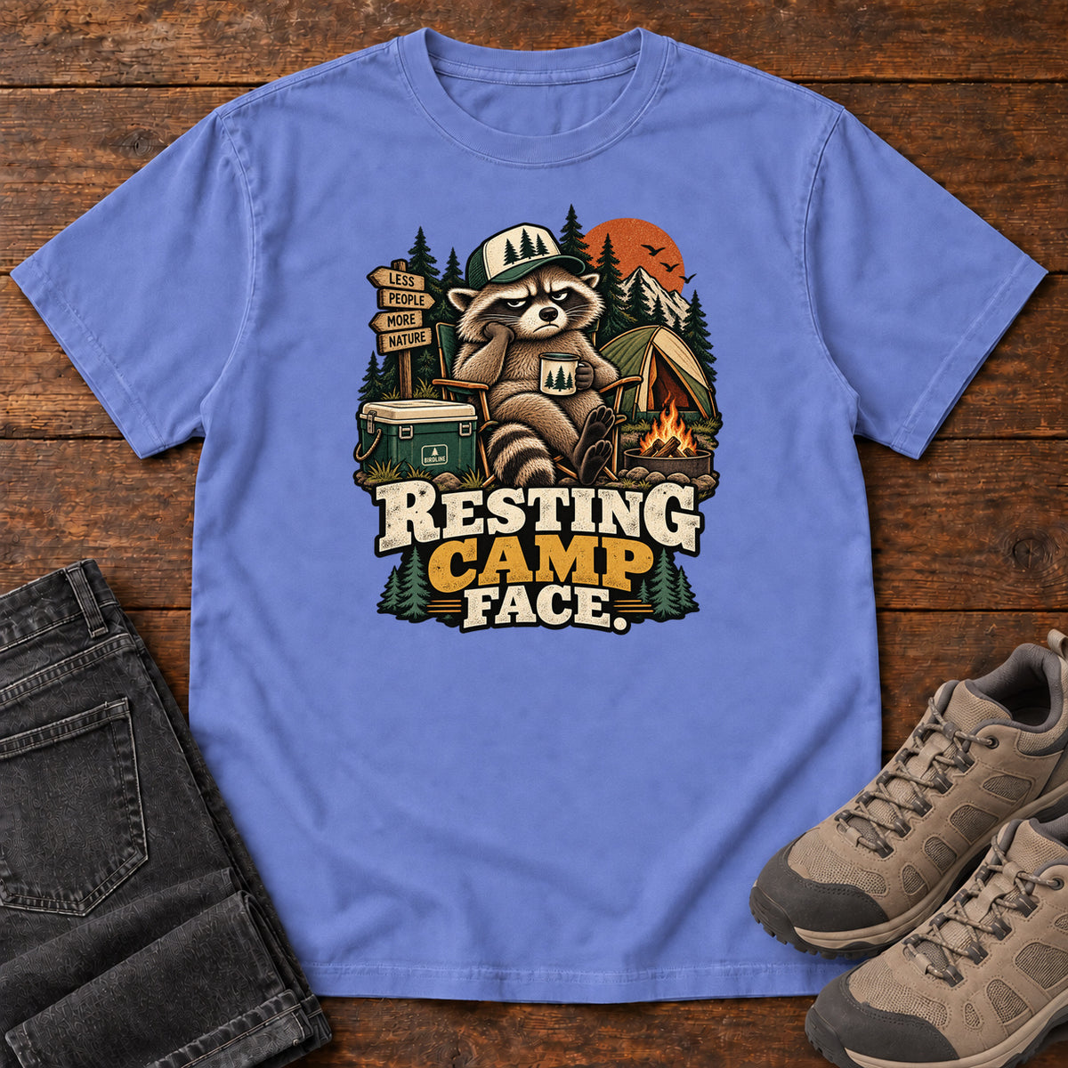 Resting Camp Face T-Shirt in Flo Blue, front laid flat on wood, featuring a capped raccoon resting in a camp chair with a mug beside a tent, cooler and campfire.
