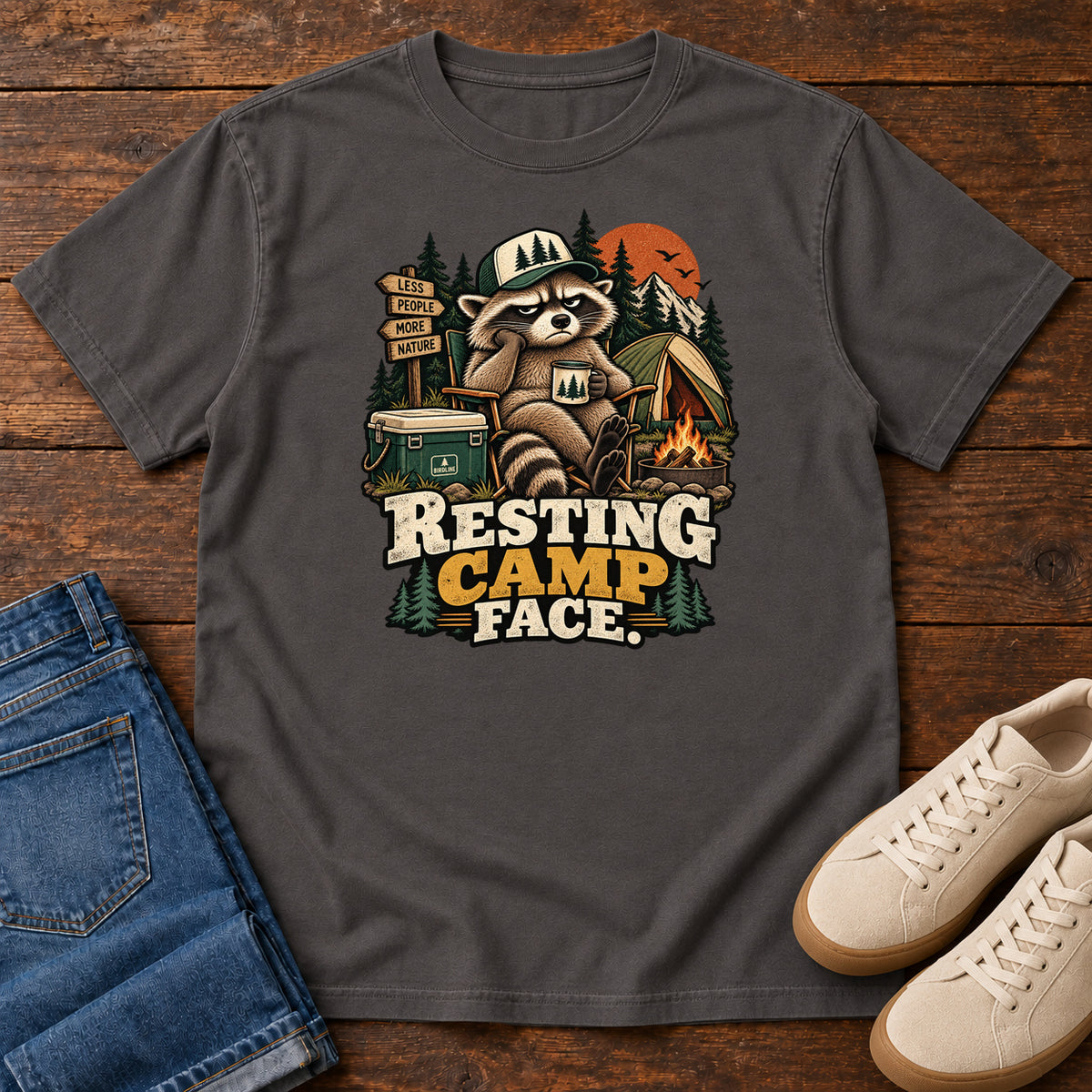 Resting Camp Face T-Shirt in Graphite, front laid flat on wood, featuring a capped raccoon resting in a camp chair with a mug beside a tent, cooler and campfire.