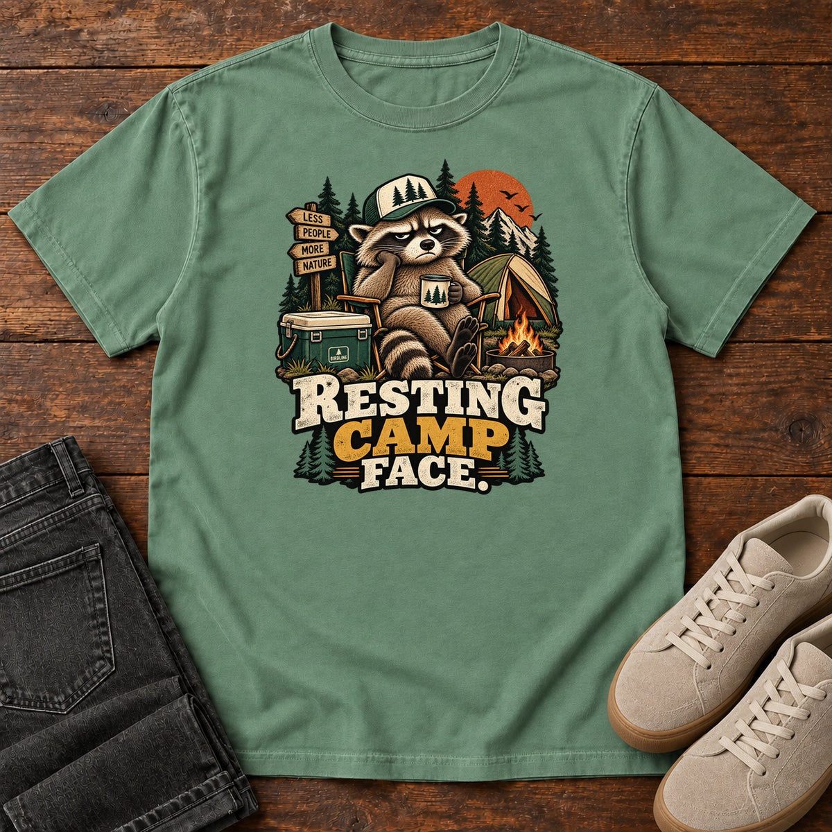 Resting Camp Face T-Shirt in Light Green, front laid flat on wood, featuring a capped raccoon resting in a camp chair with a mug beside a tent, cooler and campfire.