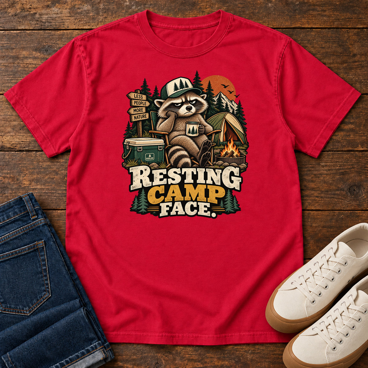 Resting Camp Face T-Shirt in Red, front laid flat on wood, featuring a capped raccoon resting in a camp chair with a mug beside a tent, cooler and campfire.