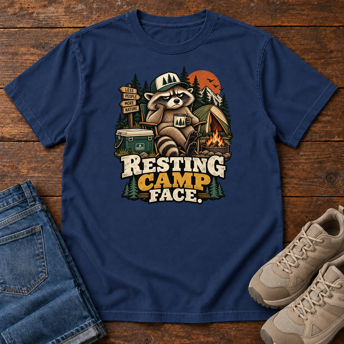 Resting Camp Face T-Shirt in True Navy, front laid flat on wood, featuring a capped raccoon resting in a camp chair with a mug beside a tent, cooler and campfire.