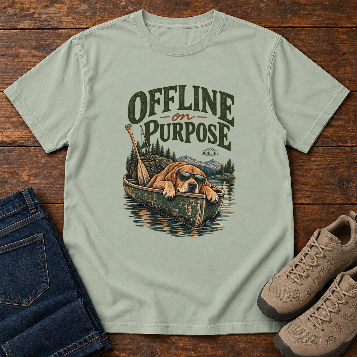 Bay Offline on Purpose Canoe T-Shirt with a brown dog wearing sunglasses and resting in a canoe below dark green lettering, front flat lay on wood.