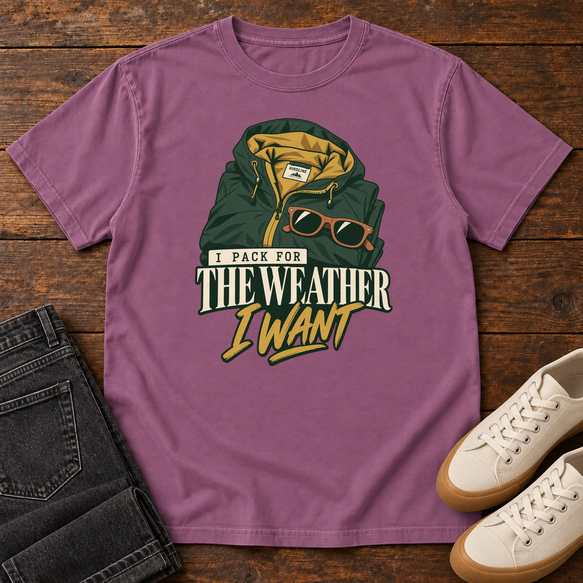 Berry I Pack for the Weather I Want T-Shirt with a green hooded jacket and sunglasses above cream and gold lettering; front flat lay on wood.