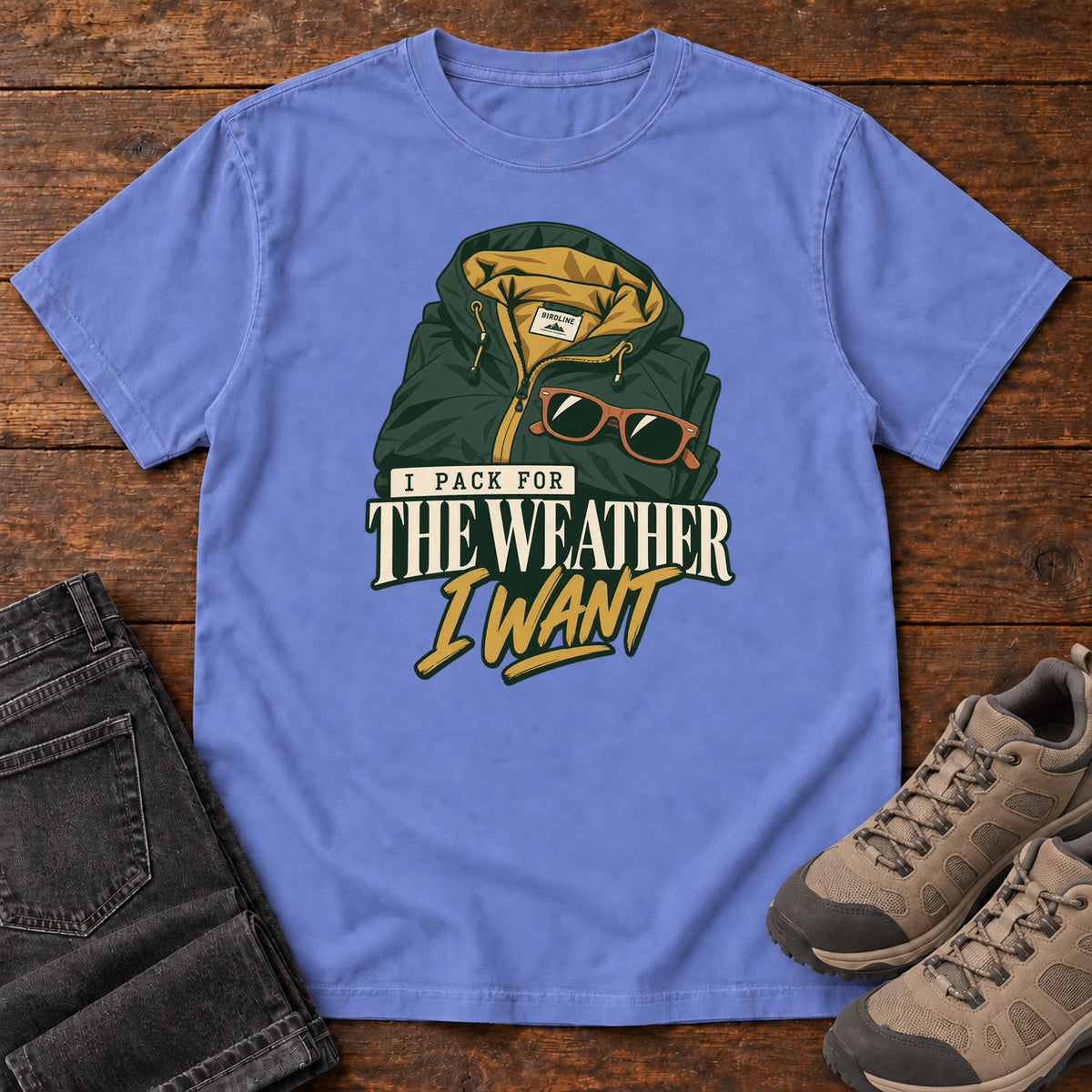 Flo Blue I Pack for the Weather I Want T-Shirt with a green hooded jacket and sunglasses above cream and gold lettering; front flat lay on wood.