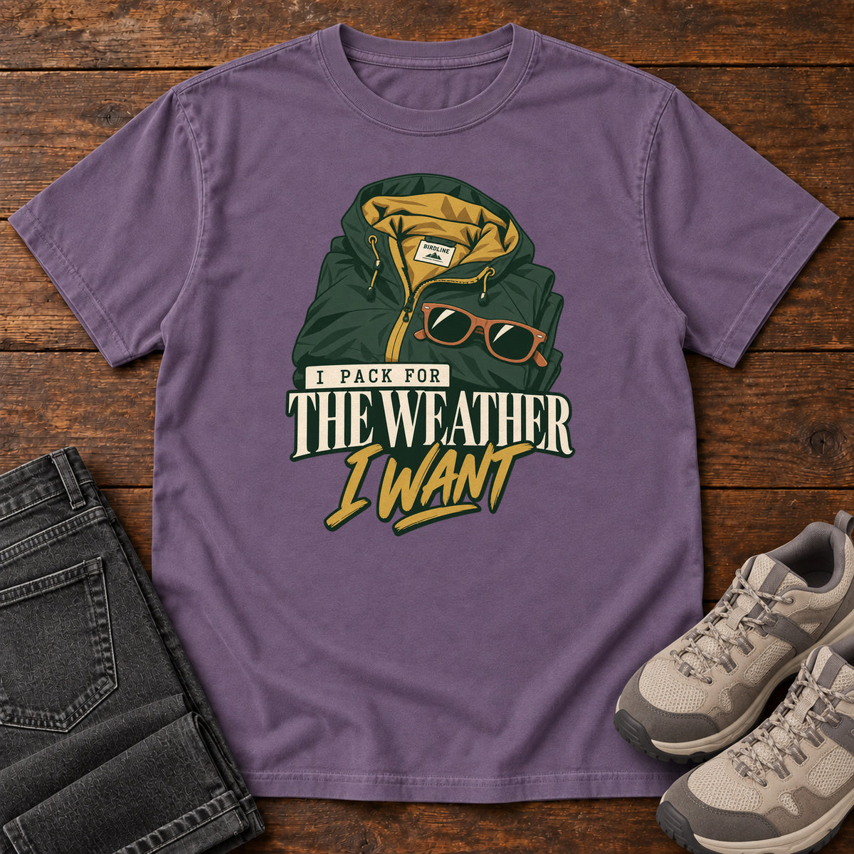 Grape I Pack for the Weather I Want T-Shirt with a green hooded jacket and sunglasses above cream and gold lettering; front flat lay on wood.
