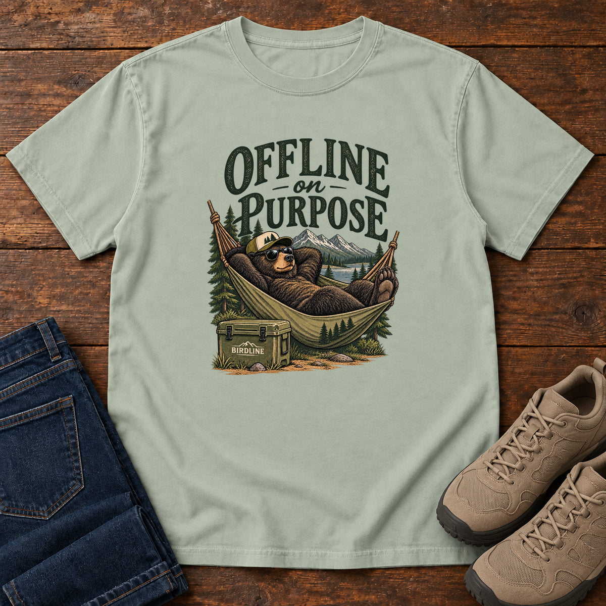 Bay Offline on Purpose T-Shirt with a bear in sunglasses relaxing in a hammock beside a cooler and mountain lake; front flat lay on wood.