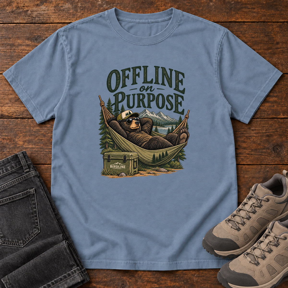 Blue Jean Offline on Purpose T-Shirt with a bear in sunglasses relaxing in a hammock beside a cooler and mountain lake; front flat lay on wood.