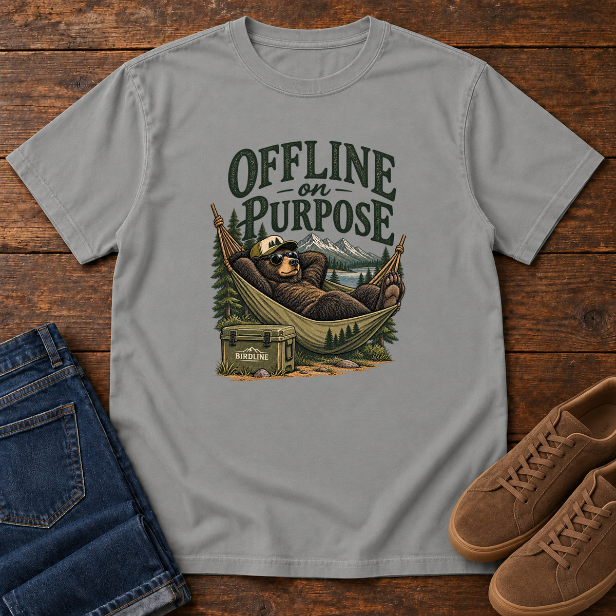 Grey Offline on Purpose T-Shirt with a bear in sunglasses relaxing in a hammock beside a cooler and mountain lake; front flat lay on wood.