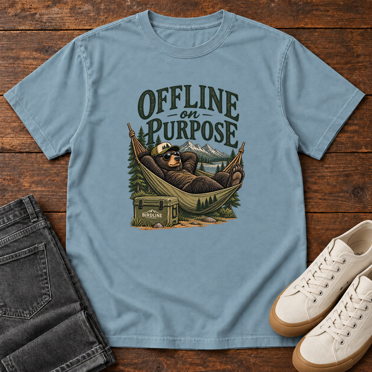 Ice Blue Offline on Purpose T-Shirt with a bear in sunglasses relaxing in a hammock beside a cooler and mountain lake; front flat lay on wood.