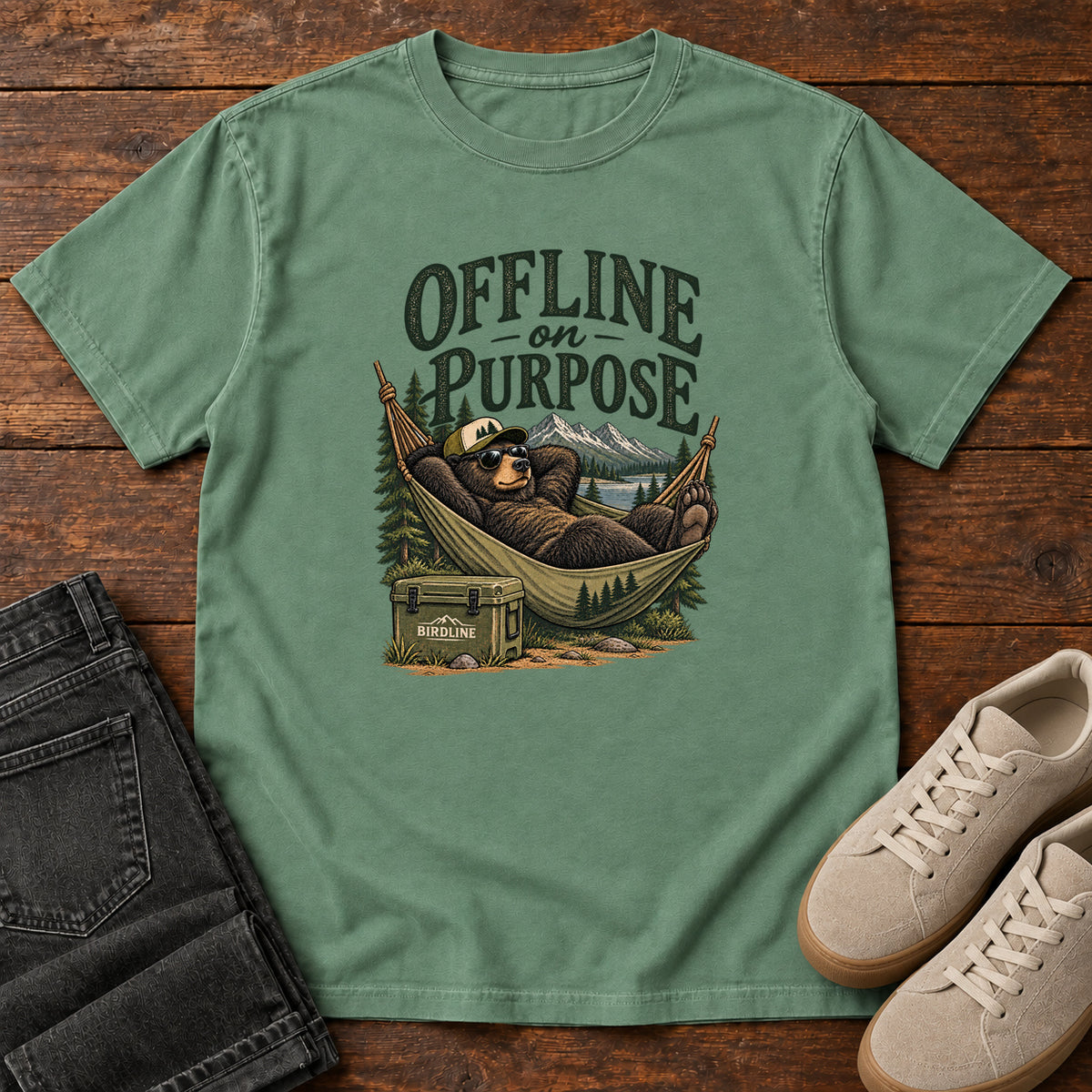 Light Green Offline on Purpose T-Shirt with a bear in sunglasses relaxing in a hammock beside a cooler and mountain lake; front flat lay on wood.