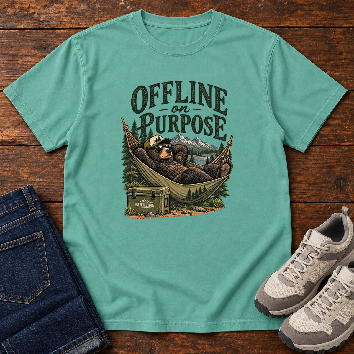 Seafoam Offline on Purpose T-Shirt with a bear in sunglasses relaxing in a hammock beside a cooler and mountain lake; front flat lay on wood.