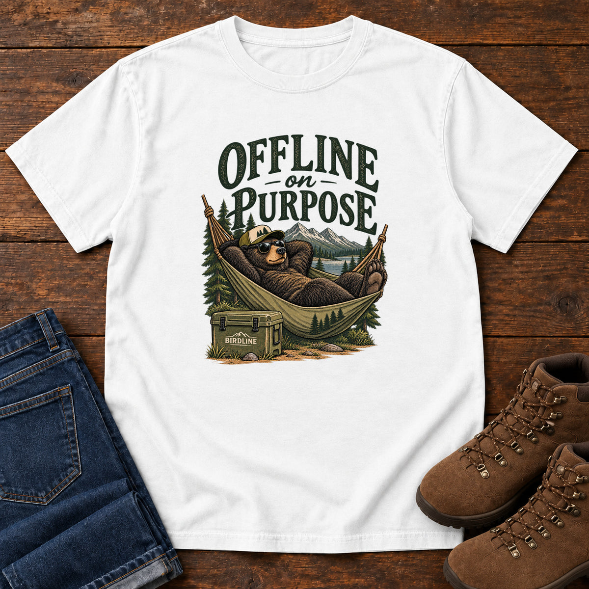 White Offline on Purpose T-Shirt with a bear in sunglasses relaxing in a hammock beside a cooler and mountain lake; front flat lay on wood.