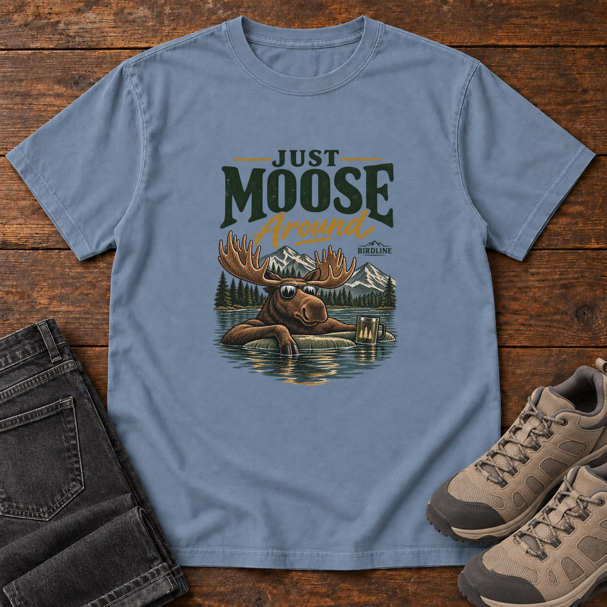 Blue Jean Just Moose Around T-Shirt with a moose wearing sunglasses in a lake beneath mountains and slogan lettering; front flat lay on wood.