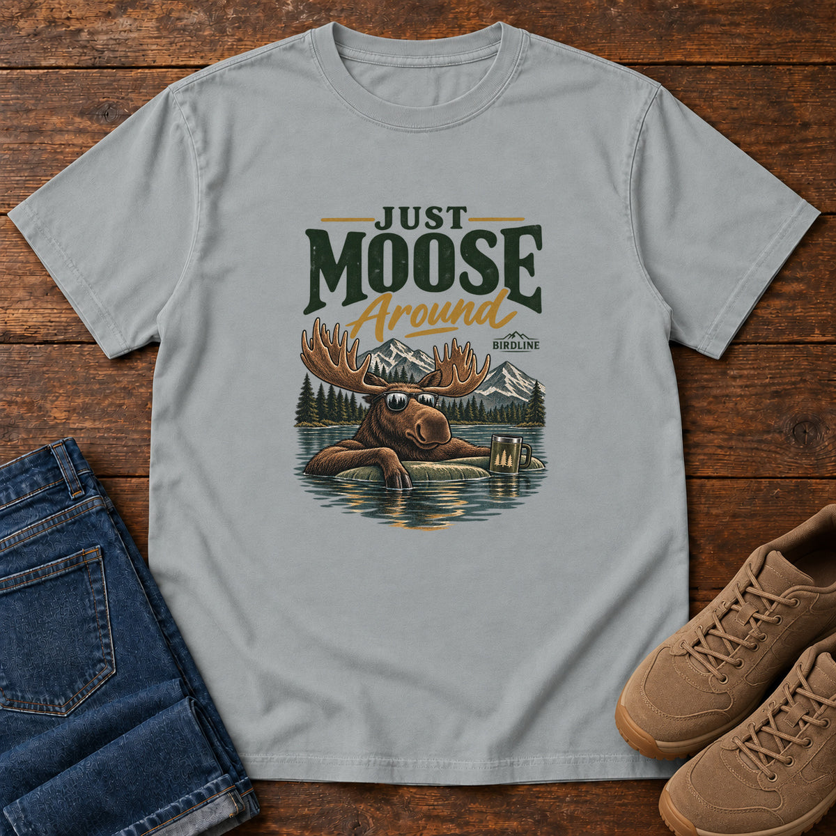 Granite Just Moose Around T-Shirt with a moose wearing sunglasses in a lake beneath mountains and slogan lettering; front flat lay on wood.